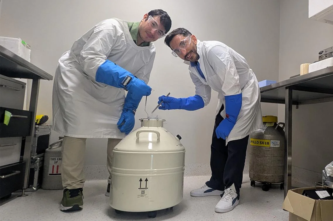 Two scientists posing with a liquid nitrogen storage cannister in a laboratory.